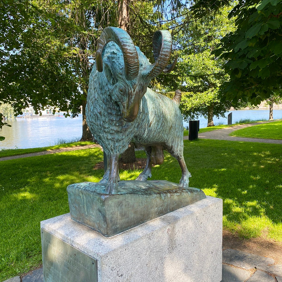 Pässipatsas Olavinlinnan puistossa. Taustalla nurmikkoa, puita ja vettä.