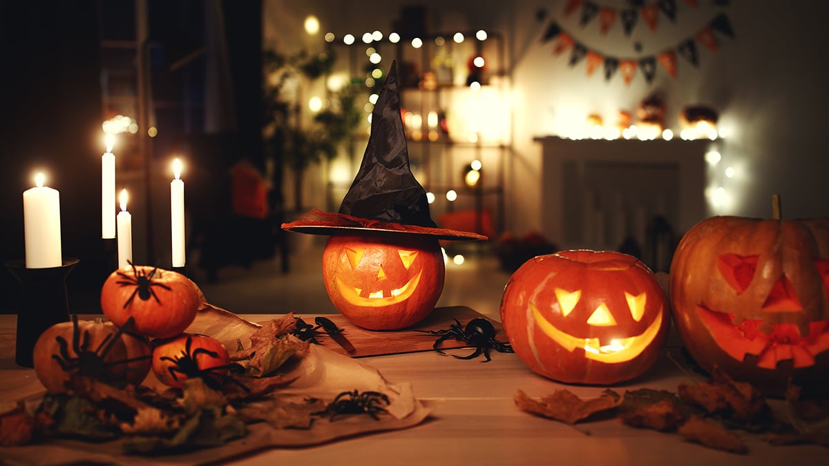 Halloween-sisustusta kotona: puisella pöydällä kynttilöitä, kurpitsoita ja hämähäkkejä. Taustalla näkyy mustaoransseja viirejä ja valonauhaa.
