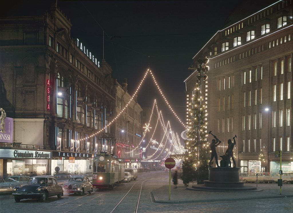 Helsingin Aleksanterinkatu hämärässä. Keskellä raitiovaunu ja autoja, oikealla Kolmen sepän patsas, taustalla jouluvaloja. Ei lunta.