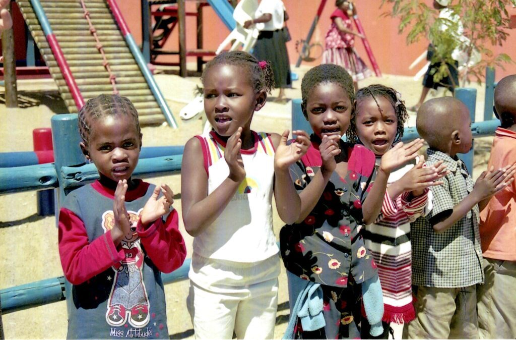 Namibialaisia lapsia Ambomaan Oshakatissa. Lapset ovat leikkimässä taputusleikkiä. Taustalla näkyy leikkipuiston välineitä.