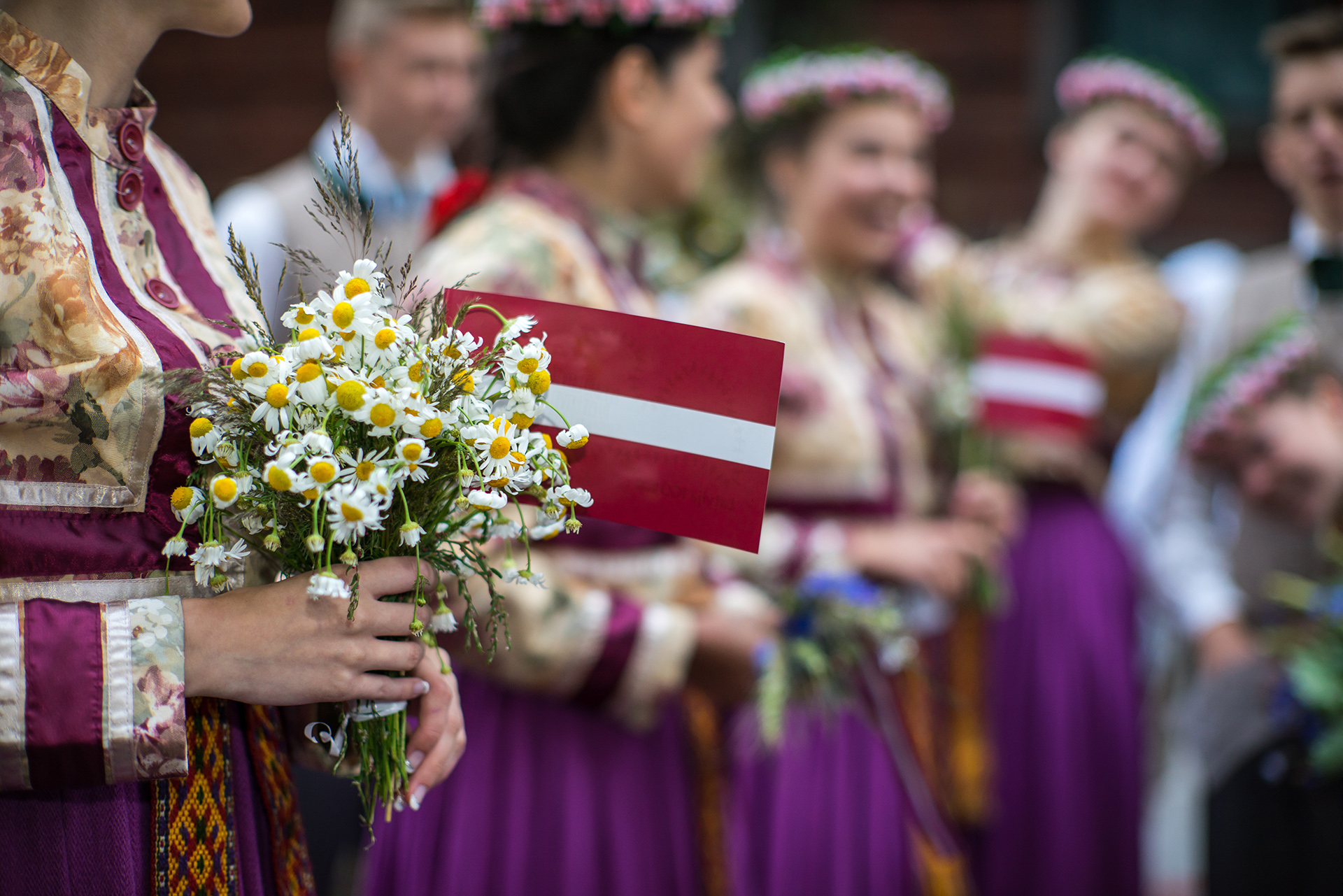 Kansallispukuisia nuoria rivissä. Nuorilla on käsissään pieniä latvian lippuja ja kukkakimppuja.
