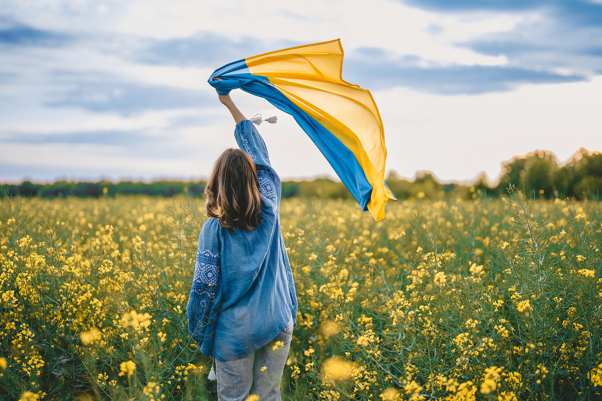 Nainen seisoo rypsipellolla selin kameraan. Naisella on sininen paita ja hän on nostanut kätensä ylös. Kädessä on Ukrainan lippu.