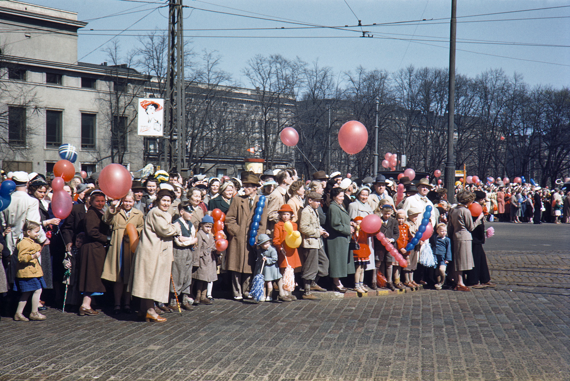 Vapunviettoa Helsingissä vuonna 1954. Kevätvaatteisiin pukeutuneet kaupunkilaiset odottavat pääsyä kadun yli. Joillakin on ylioppilaslakit ja joillakin ilmapallot.